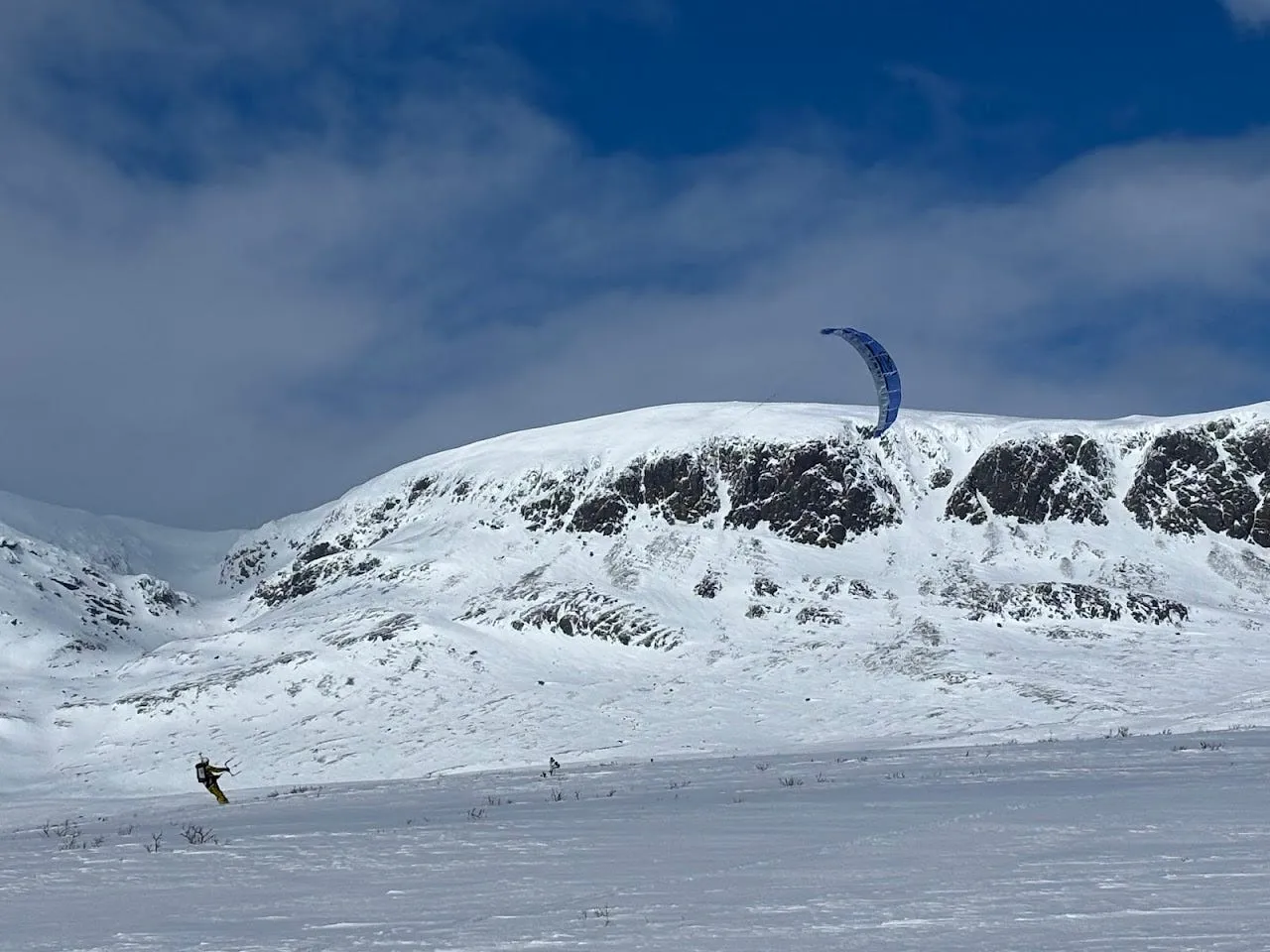 A lone snowkiter crossing an open snowfield beneath a steep, exposed mountain face under a clear sky.